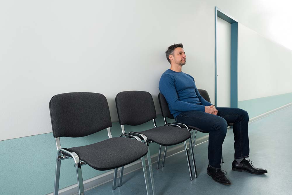 follow-up hoofd-halskanker man met kort grijs haar en blauwe trui wacht op een stoel in een ziekenhuisgang. Naast hem staan nog 2 lege stoelen.